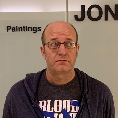 Photo of Jon Reischl, a balding man with glasses wearing a blue hoodie over a black Blood Shot Records tee shirt. There are strips of blue masking tape on his shirt. He is standing in front of a wall with the words 'paintings' and 'Jon' in vinyl lettering.